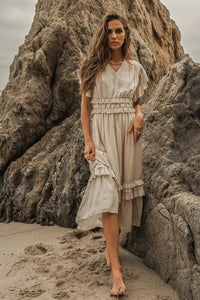 A woman on a beach next to some rocks walking bare foot wearing an ivory willa ruffle dress.