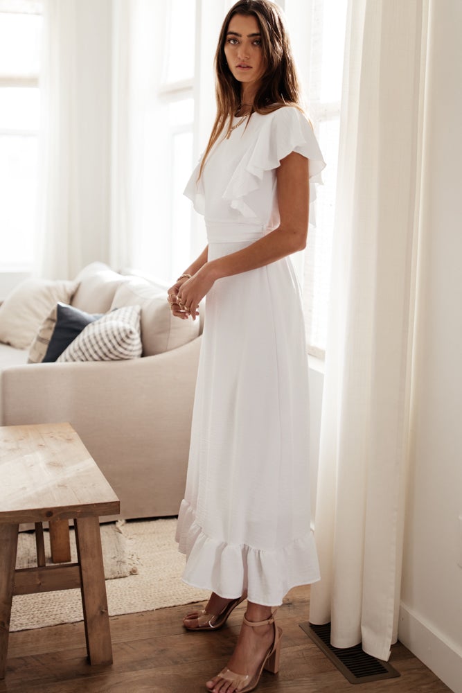 Model is standing in front of a window wearing an elegant white maxi dress with ruffled sleeves and a tied waist.
