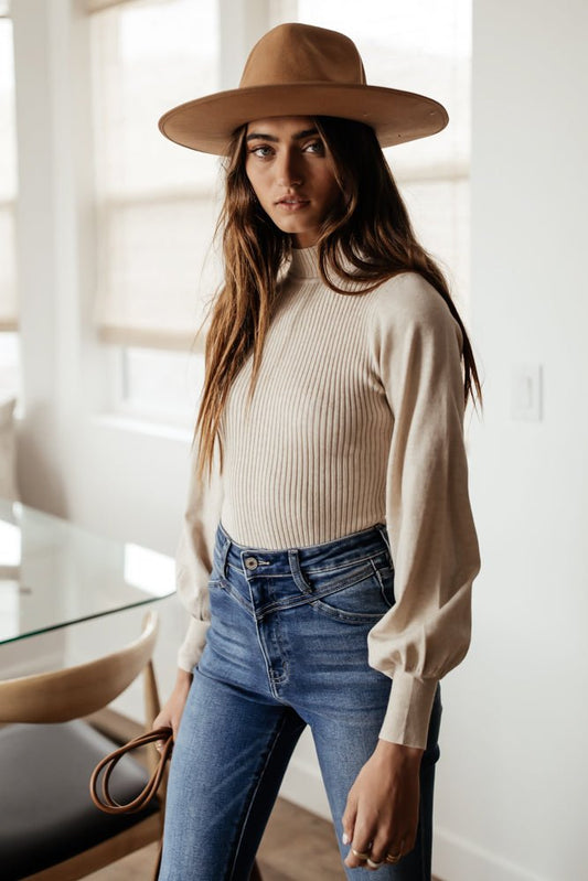 Model wears the Emily Ribbed Bodysuit in Cream with medium wash jeans and a tan hat. Bodysuit has mock neck and tapered sleeves.