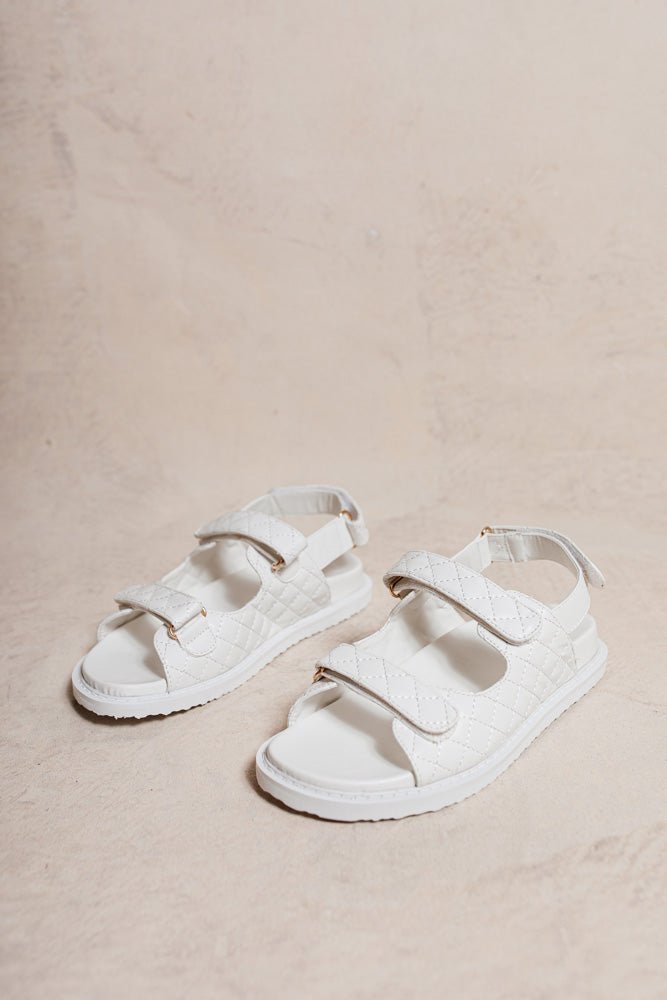 The Rory Sandals in White have two velcro straps across the top, back ankle strap with velcro, gold hardware, and criss cross stitching. 