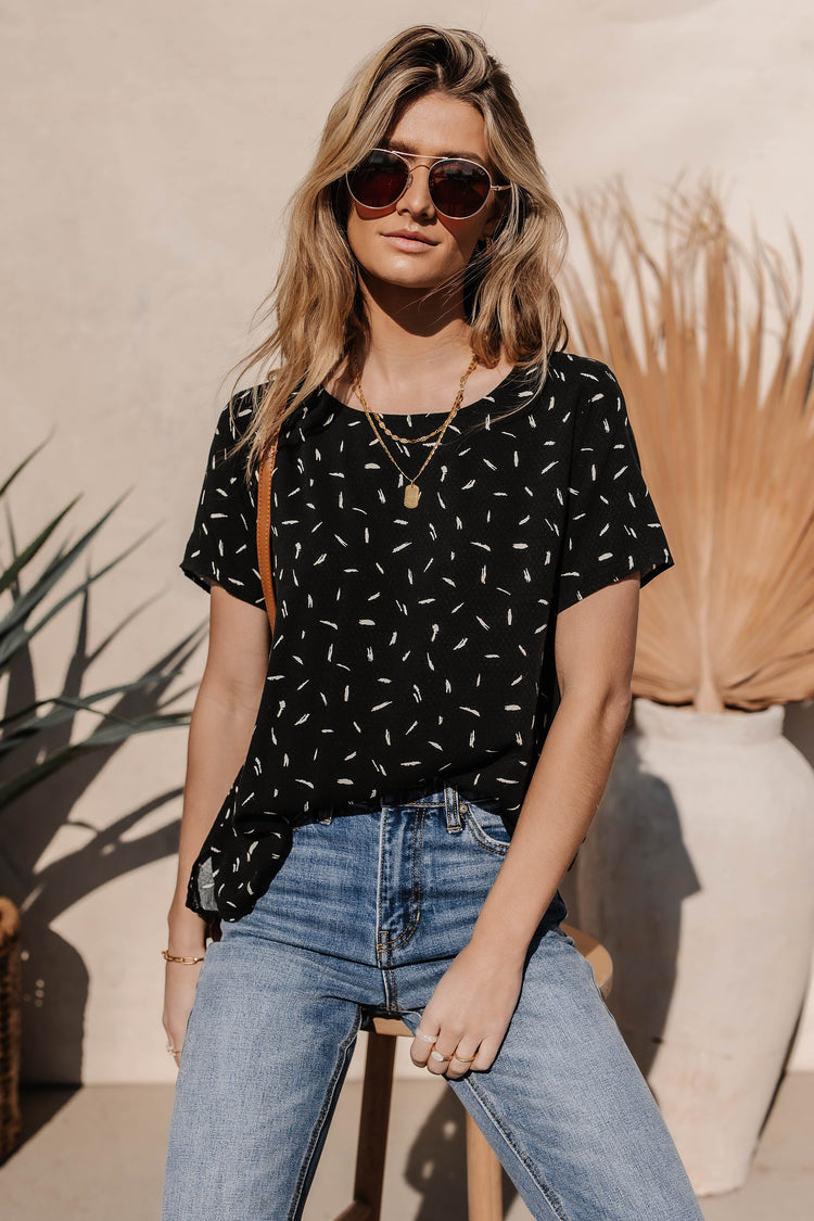 Model wears the Victoria Shirt in Black with medium wash jeans, gold necklaces, and brown sunglasses. Shirt has relaxed fit and abstract pattern.
