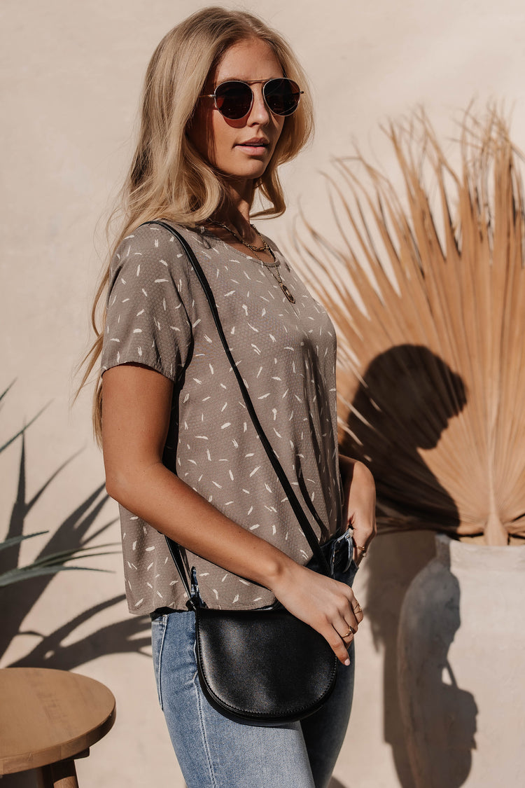 Model wears the Victoria Shirt in Taupe with medium wash jeans, a black purse, and brown sunglasses. Top has short sleeves and relaxed fit.