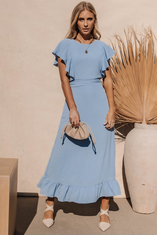 Model wears the Janet Ruffle Sleeve Maxi Dress in Light Blue with white heels and a blush clutch. Dress has ruffle detail on hem and shoulders.