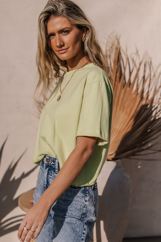 Model wears the Jane Cropped Tee Shirt in Green with light wash dneim and a gold necklace. Tee has relaxed fit and raw hem.
