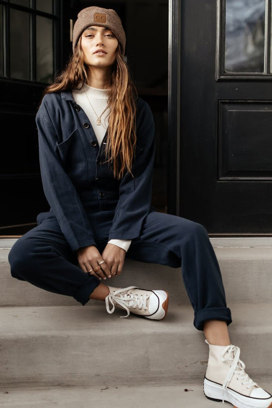 Model wears the Ellwood Jumpsuit in navy with a white top, cream platform sneakers, and a brown beanie. Jumpsuit has button front, long sleeves, and rolled leg.