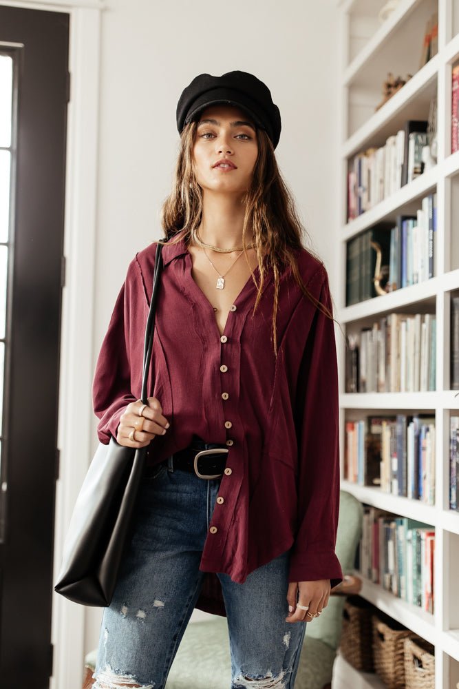Model wears the Rosealie Oversized Shirt in Burgundy with medium wash skinny jeans, black cap, and black belt. Shirt has button front and v-neck.