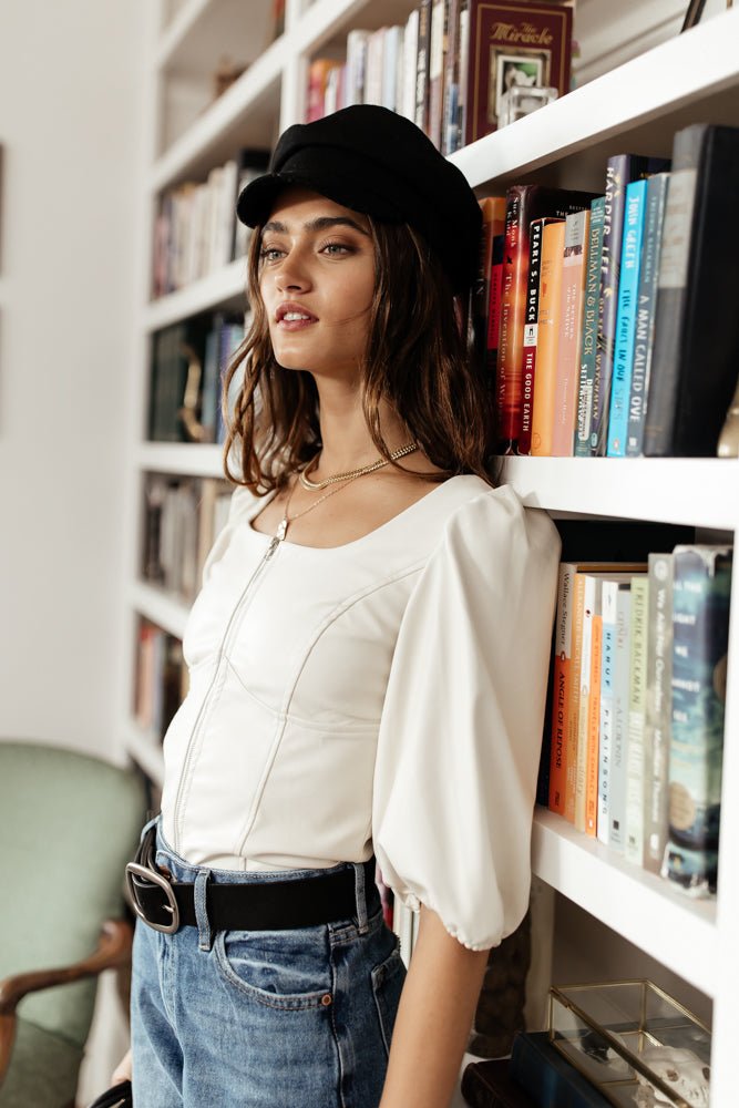 Model wears the Makena Vegan Leather Top with medium wash jeans, a black cap, and black belt. Top has puff sleeves and a zipper front.