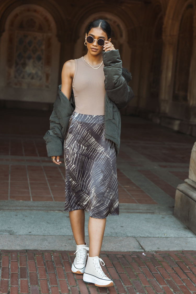 Model wears the Fleur Midi Skirt with a nude tank top, green jacket, and cream platform sneakers. Skirt has white abstract stripe pattern.