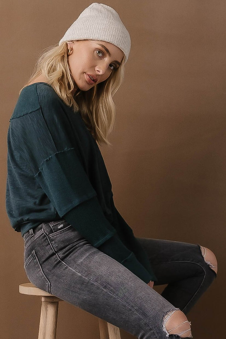 A woman sitting on a stool and looking sideways while wearing a Galilea patch top in teal.