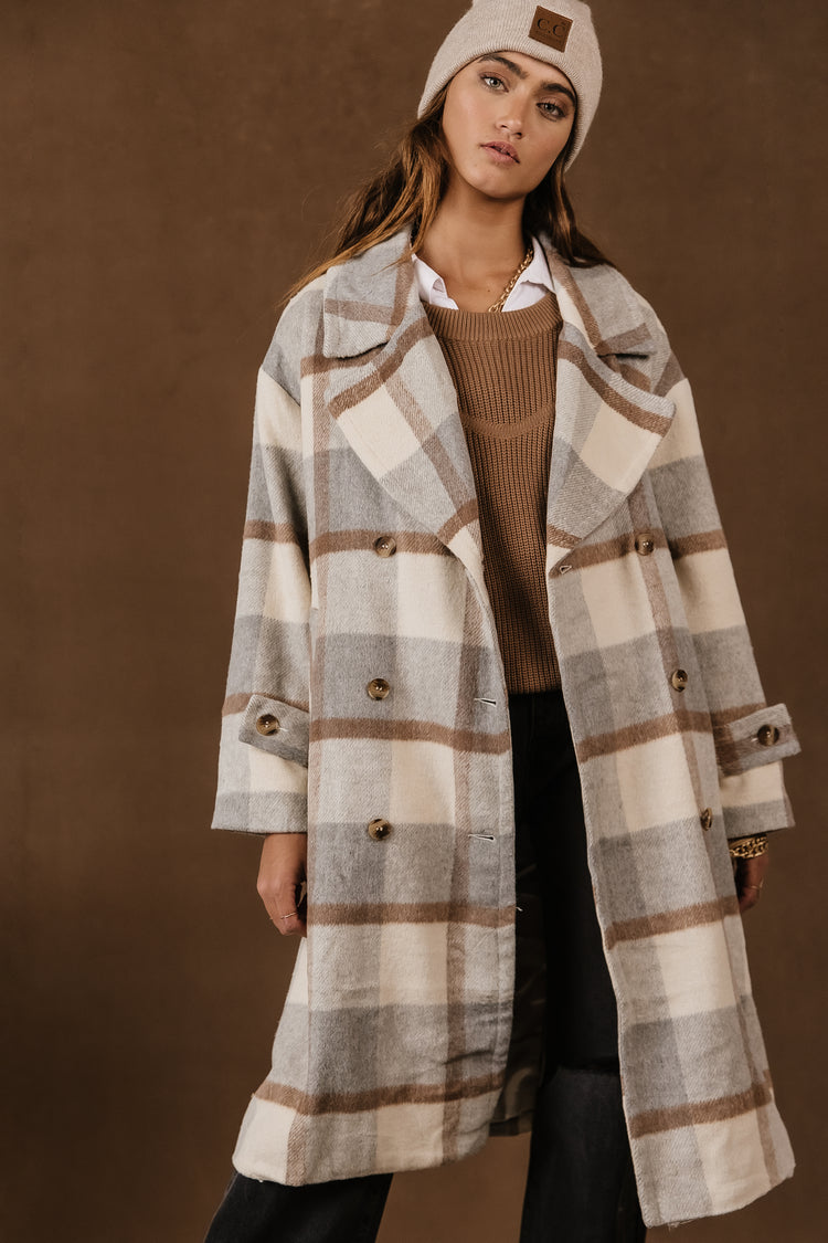 Model wears the Casper Trench Coat with a tan sweater, black jeans, and a beige beanie. Coat has brown, grey, and cream plaid pattern.