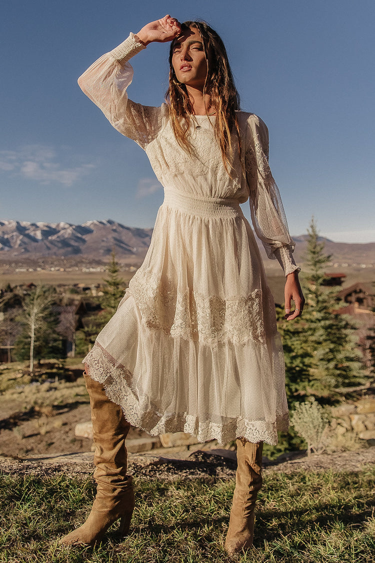 Model wears the Lilith Midi Dress with taupe suede boots. Dress has smocked waist, long sleeves, and lace details.