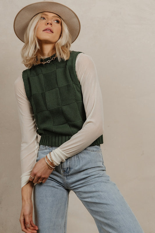 Model wears the Chloe Sweater Vest in Emerald with light wash denim, cream long sleeve shirt, and a beige hat. Vest has checker knit pattern.