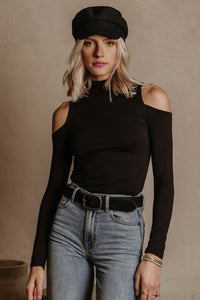Model wears the Cathy Open Shoulder Top in Black with light wash jeans, a black paperboy cap, and a black belt. Top has turtleneck and shoulder cut outs.