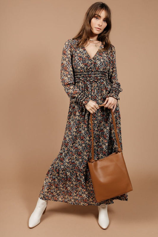 Model wears the Angelica Maxi Dress with white heeled boots and a brown purse. Dress has v-neck, smocked cuffs, and empire waistline.