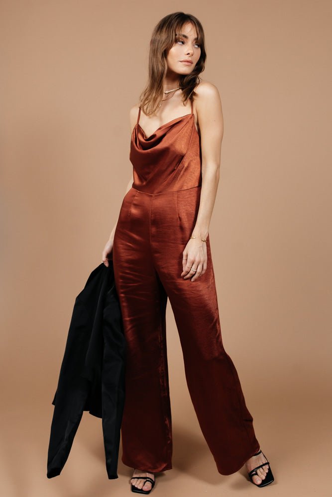Model wears the Agna Jumpsuit in Cognac with strappy black heels. Jumpsuit has a cowl neck, long length, and spaghetti straps.