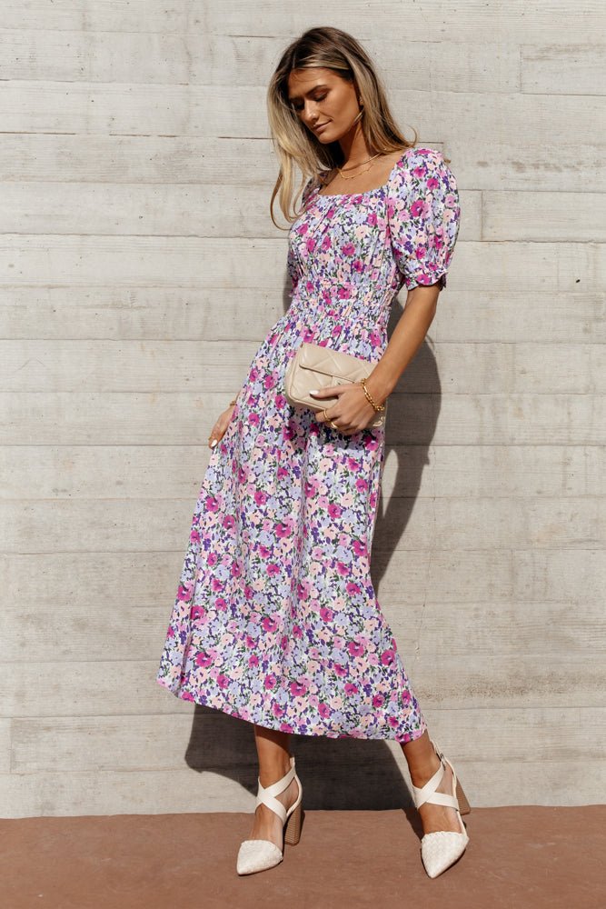 Model wears the Keely Floral Dress with a blush clutch and white heels. Dress has puff sleeves, elasticized waist, and midi length.
