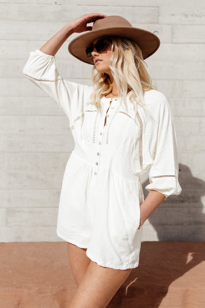 Model wears the Campbell Romper with a brown hat and brown sunglasses. Romper has 3/4 sleeves, button front, and high neck.