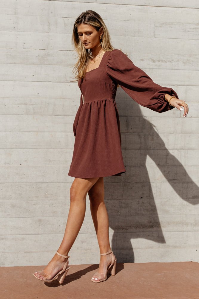 Model wears the Alma Mini Dress in Brown with nude heels. Dress has square neck, smocked back, and balloon sleeves.