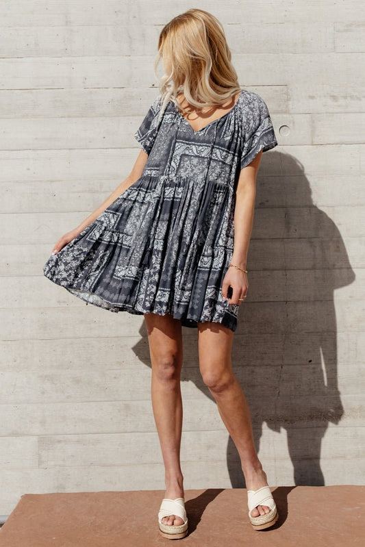 A woman with her hair over her face as she is looking to the left and she is standing in front of a textured wall and she is wearing a floral mini dress in charcoal.