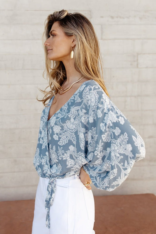Model wears the Sydney Wrap Top with white denim and gold jewelry. Top has balloon sleeves, v-neck, and floral pattern.