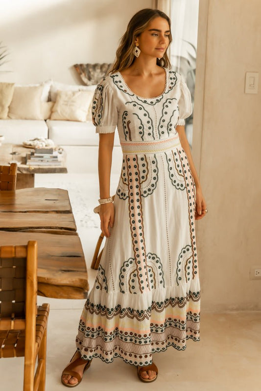 Model wears the Avalee Maxi Dress with brown strap sandals and gold earrings. Dress has round neck, short puff sleeves, and colorful embroidery.