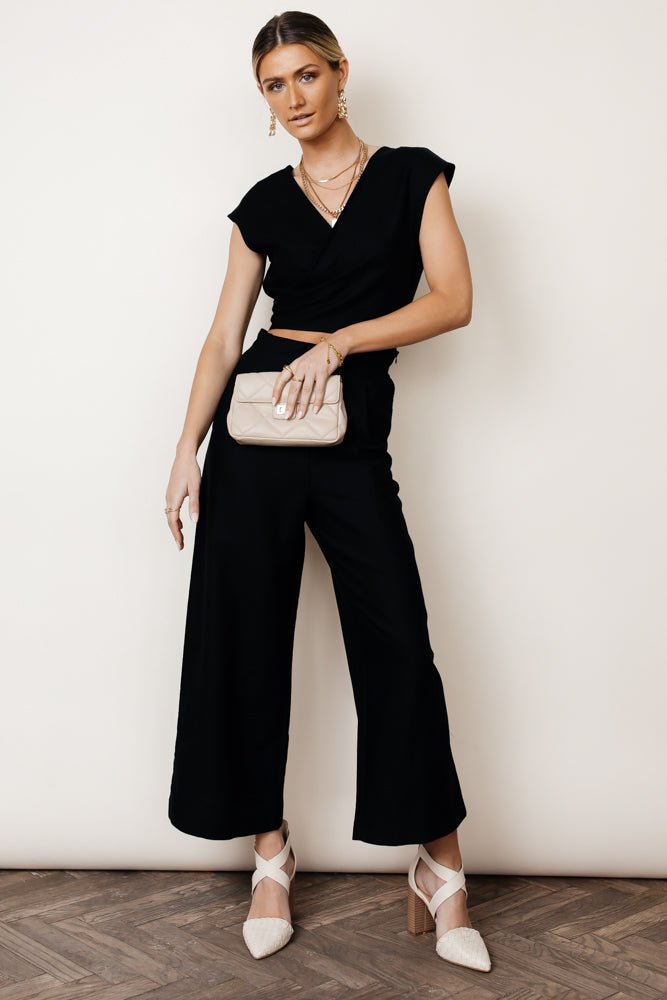 Model wears the Lua Wrap Jumpsuit in Black with white heels and a beige clutch. Jumpsuit has v-neck, wide leg, and waist cut-out.