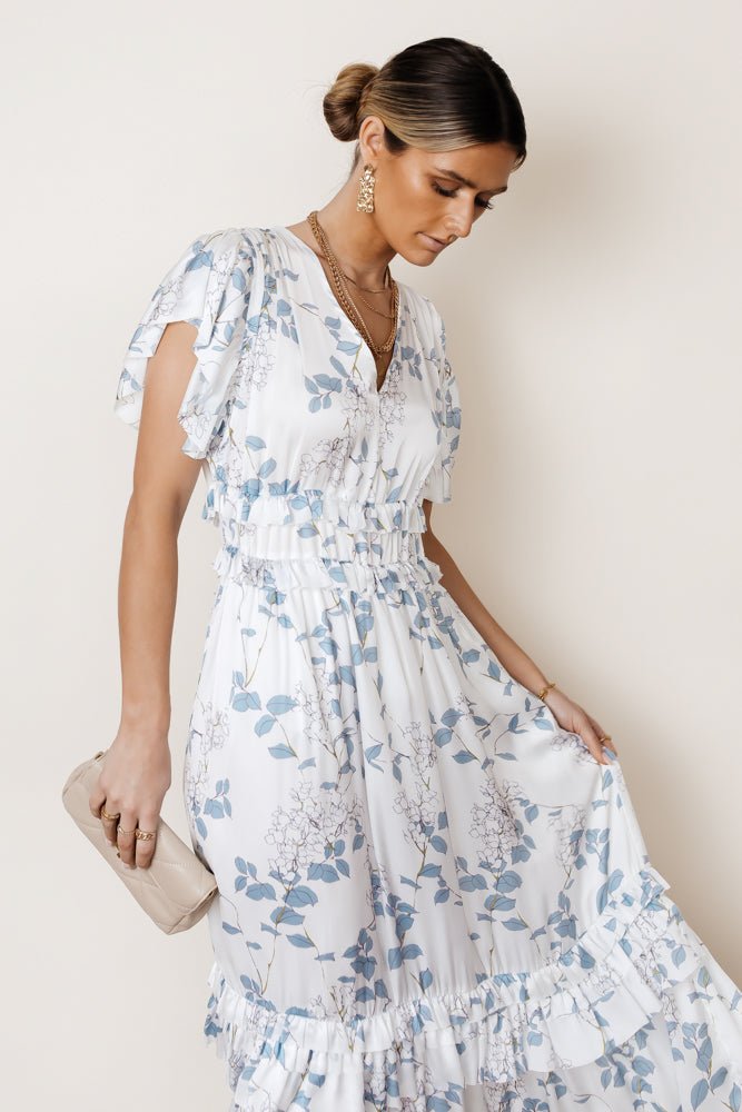 Model wears the Willa Ruffle Dress in Blue Floral with a blush clutch and gold jewelry. Dress has v-neck, flutter sleeves, and ruffle details on waist and skirt.