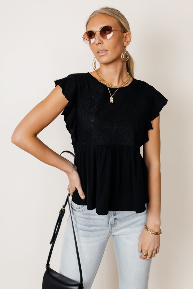Model wears the Catalina Peplum Top in Black with light wash jeans, sunglasses, and a black purse. Top has flutter sleeves and peplum.