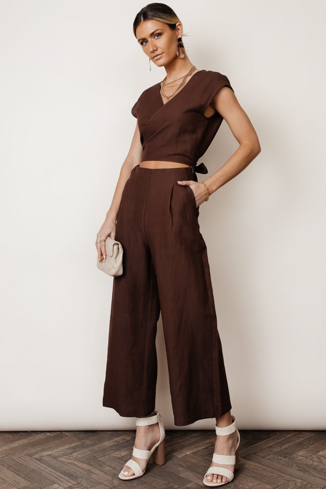Model wears the Lua Wrap Jumpsuit in Brown with white heels and a beige clutch. Jumpsuit has waist cutout, v-neck, and wide leg.