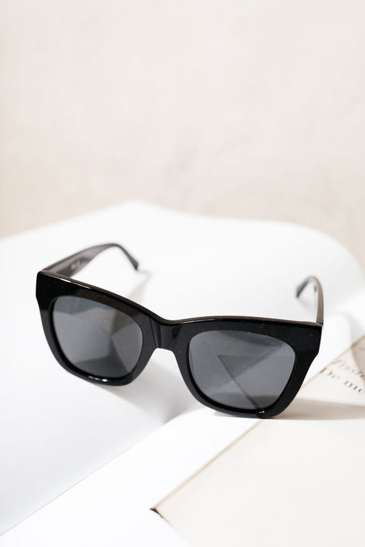 The Billie Sunglasses in Black has thick black frames and black tinted lenses. Sunglasses are displayed on an open book.