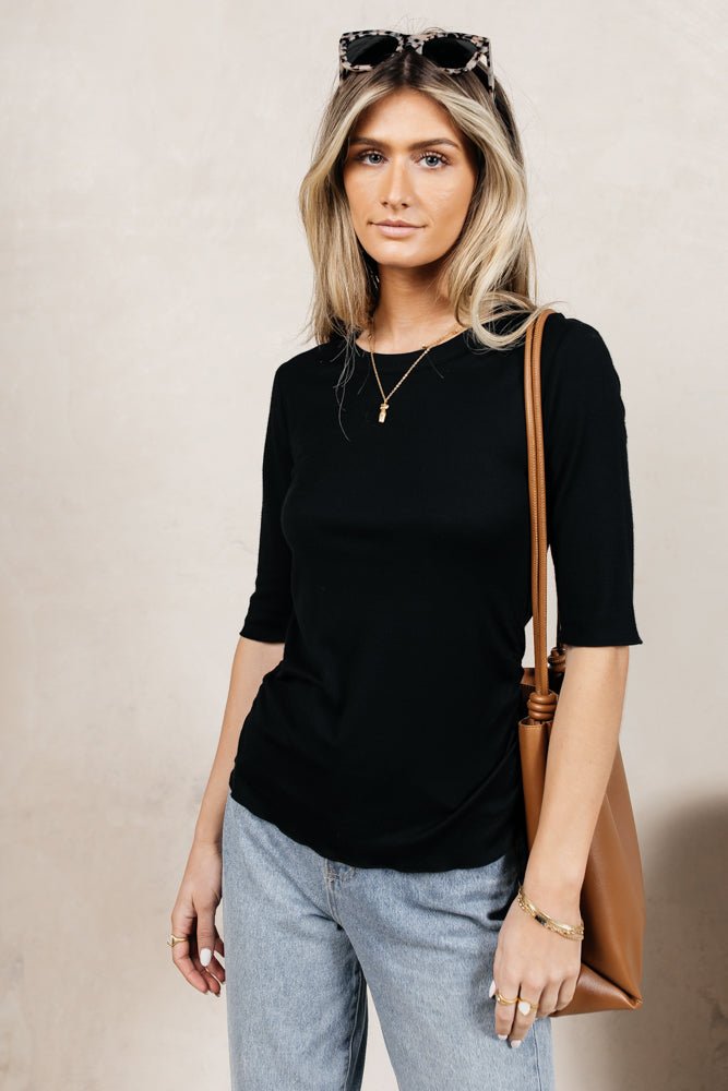 Model wears the Sutton Ribbed Top in Black with light wash denim, sunglasses, and a brown purse. Top has ribbed material, crew neck, and elbow length sleeves.