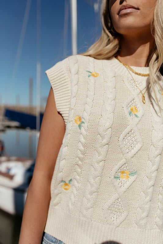 Model wears the Calla Sweater Vest with gold necklaces and light wash denim. Vest has high neck, cable knit, and small embroidered yellow flowers.