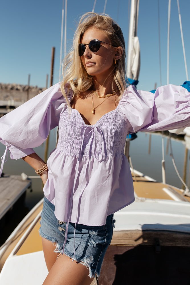 Model wears the Cheralyn Blouse in Lavender with denim cutoff shorts and sunglasses. Blouse has smocked bodice, balloon sleeves and tie detail.