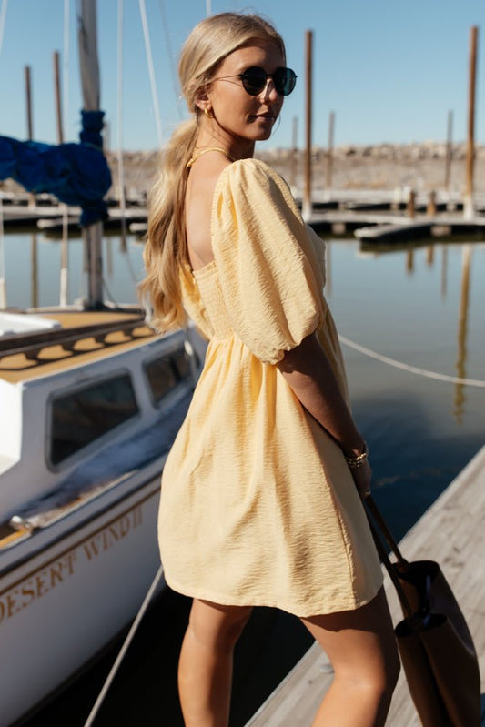 A woman standing on a dock next to a sail boat and she has a ponytail and is wearing a yellow mini dress with puff sleeves.