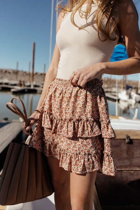 Model wears the Karrington Mini skirt with a cream tank top and tan purse. Skirt has elastic waist, layered details, and ditsy floral pattern.