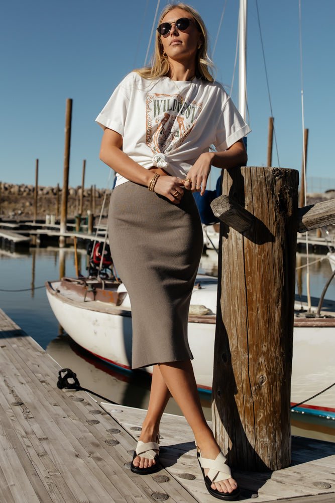 Model wears the Roisin Midi skirt in Sage with a white graphic tee, white sandals, and sunglasses. Skirt is fitted and reaches model's knees.