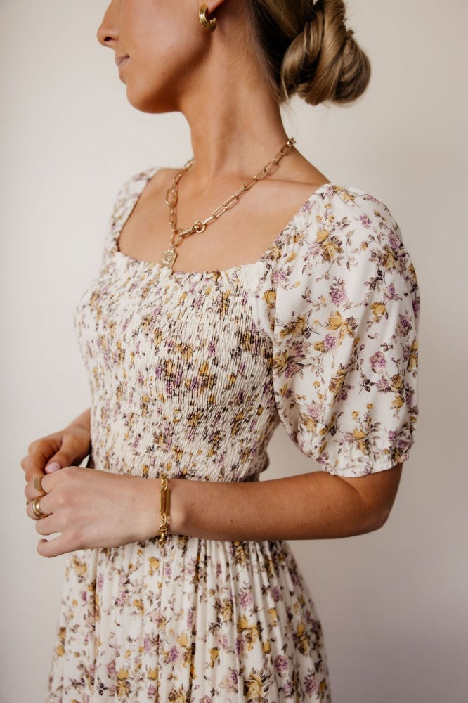 Model is shown from the head to the waist wearing a puff-sleeve floral dress with a white base and multi-colored flowers, and a fitted waist.