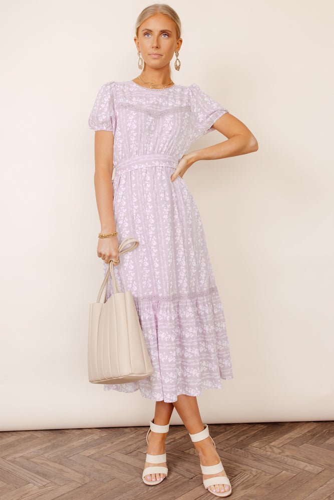 Woman with her left hand on her hip wearing big earrings, a lilac midi dress and heels.
