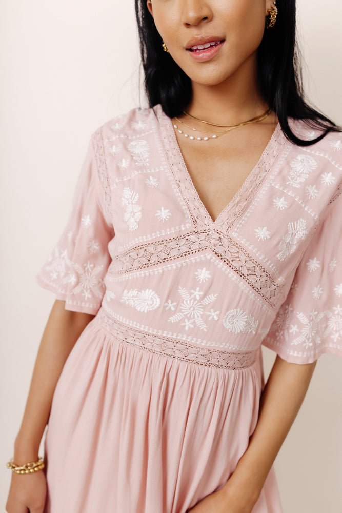 A close up photo of a woman with long black hair standing in front of an off white wall wearing the Annette Mini Dress in Blush, which has lace see through detail on the bodice, short flutter sleeves and a v-neck. She is also wearing gold and pearl jewelry. 