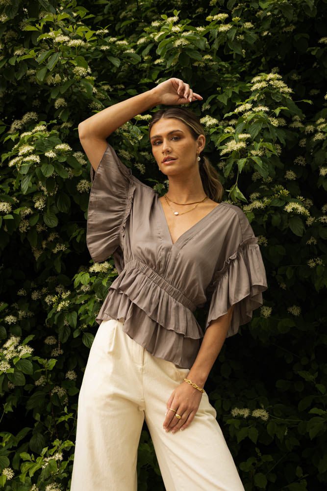 A woman in front of a tree wearing a flutter sleeves blouse in grey.