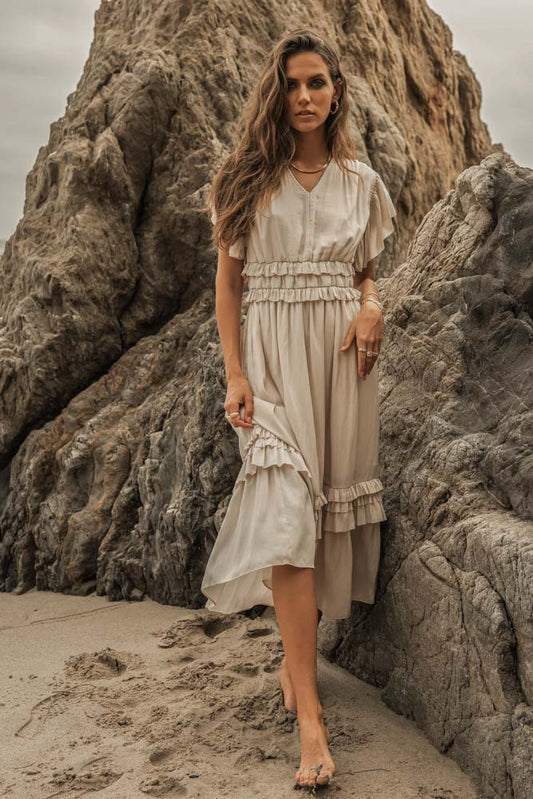 A woman on a beach next to some rocks walking bare foot wearing an ivory willa ruffle dress.